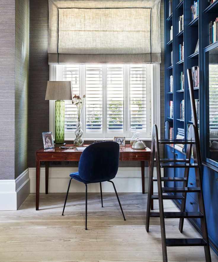 Soothing home office with cobalt blue built-in bookshelves and black wooden step ladder. The desk space is facing a window with shutters, and a linen blind at the top