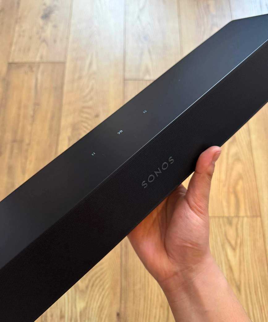 Hand holding the Sonos Ray soundbar with wooden floor in background.