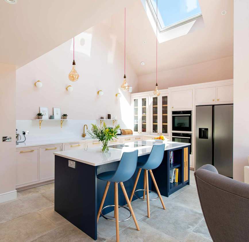 Blue kitchen island and bar stools with blush pink walls, white countertops and pale gray stone flooring.