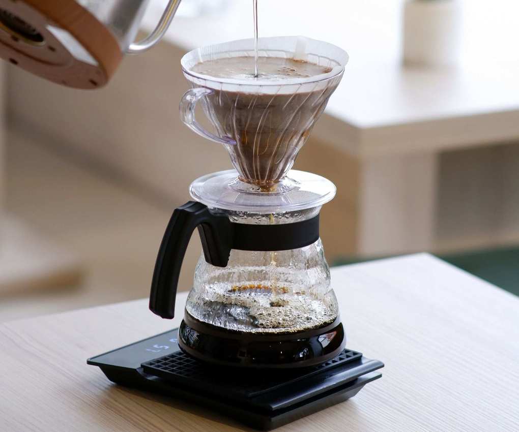 A pour over coffee maker with a glass carafe on a wooden table with a gooseneck kettle pouring into it