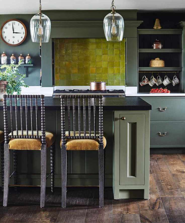 A green kitchen with brown wooden flooring, green cabinets, green tiling, and a green kitchen island with a black marble countertop. In the foreground are two wooden chairs with velvet upholstery, with two glass pendant lamps hanging above. To the right is alcove shelving with mugs and to the left is a wooden circular wall clock.