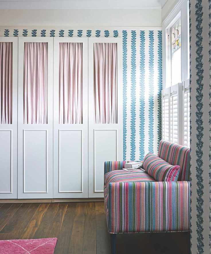 A bright and patterned bedroom with wooden flooring, pink rug, striped sofa, white and blue patterned wallpaper and a white closet with pink curtains. Large windows with white shutters flood the room with light.
