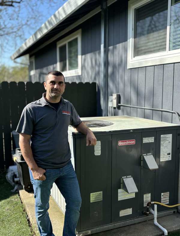 Alex Atkinson, HVAC and plumbing expert at Super Brothers, stands next to a large outdoor pump unit. He's white, with short brown shaved hair, and he is wearing a pair of blue jeans and his gray walk polo top.