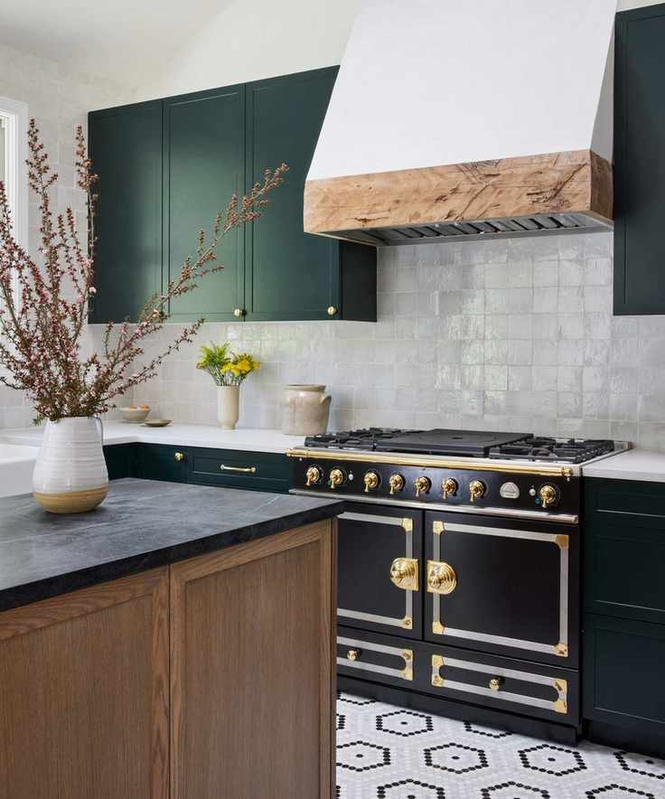 A kitchen with a Le Cornue range in black and brass, green cabinets, a reclaimed oak hood, and a backsplash made of zellige tiles.