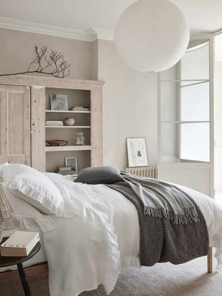 white and neutral bedroom with limed cabinet, stone walls, white bedding, cream carpet, gray blanket and cushion, white paper lampshade