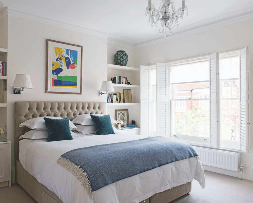 A large double bedroom with a buttoned headboard bed with white bedding, a blue blanket and blue cushions, white walls with colorful framed artwork above the bed, shelves with miscellaneous items such as books, framed pictures and vases, and large windows with shutters. A glass chandelier hangs above