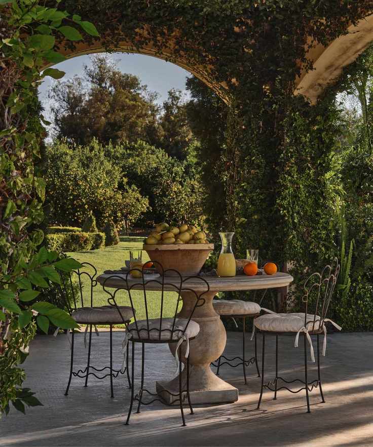 european style outdoor patio with loads of climbing green plants, a round pedestal concrete table with scalloped wrought iron chairs and a bowl of lemons