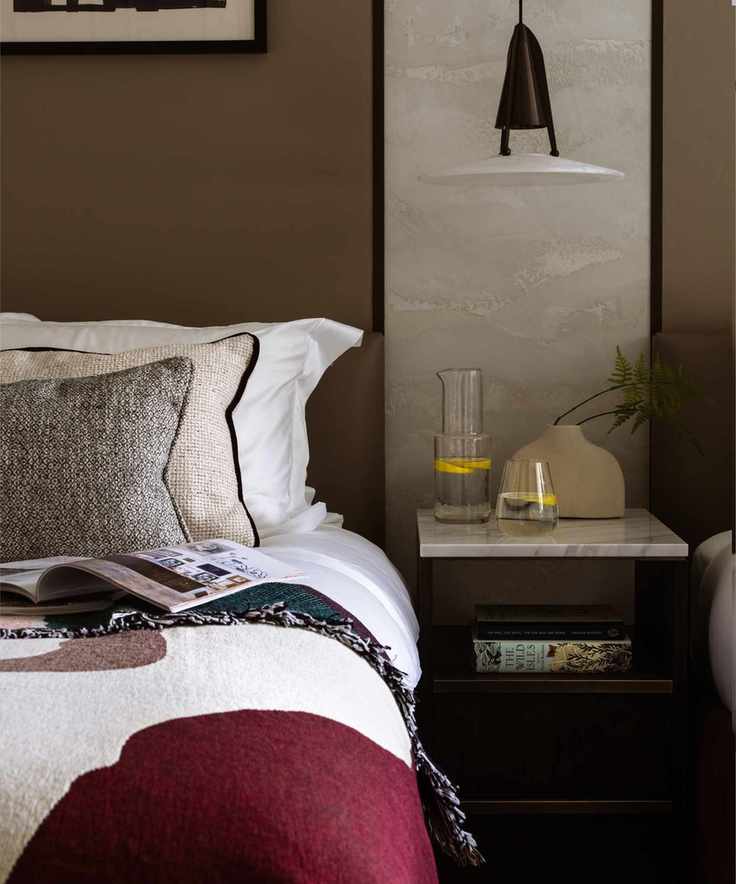 White and red bedding with gray cushions on a bed against a brown wall beside a wood nightstand.