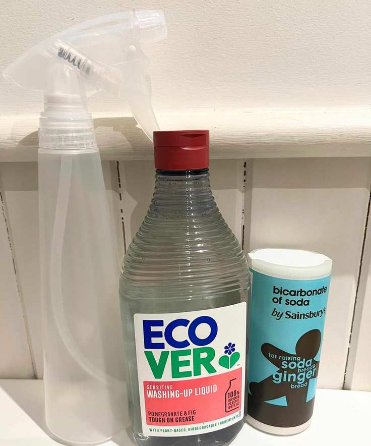 An empty clear spray bottle, a clear bottle of washing up liquid, and a blue container of baking soda with a gingerbread man on it against a white panelled wall