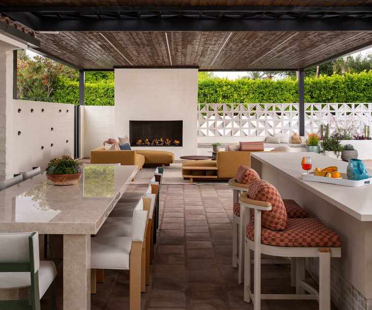 An outdoor dining and living area under cover with high chairs at a bar, a dining table and a living area with fireplace beyond
