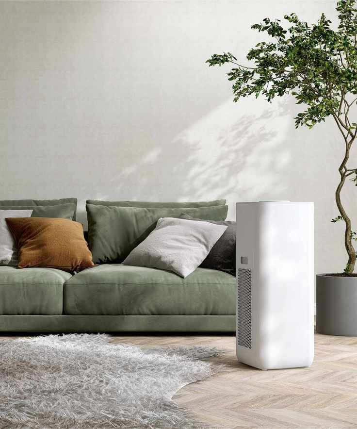 A white air purifier situated beside a green sofa and an indoor tree, with a white wall and wooden flooring in the background.