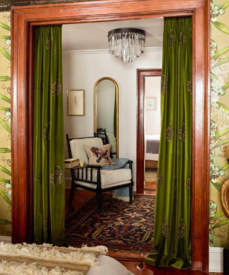 Bedroom with wood door frame, wallpapered wall, green velvet curtains looking into living area with antique wood chair and rug and glass chandelier