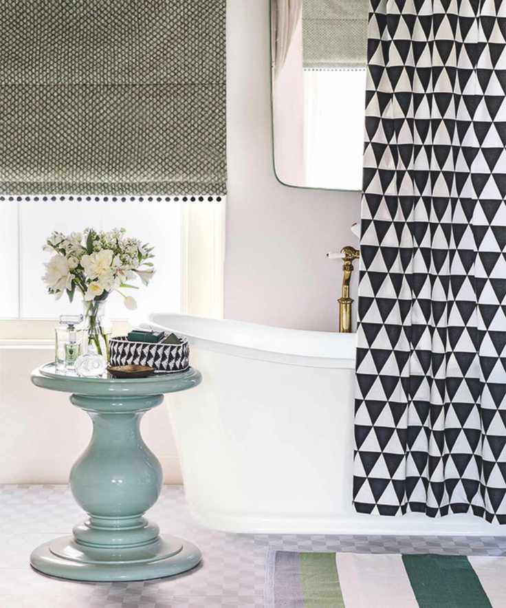 A white bathtub in a bright bathroom with a black and white shower curtain and gold faucet, mirror above, and a blue circular table with a clear vase of white flowers on top. Behind is a large window with green blind, and on the floor is a green and white striped rug.