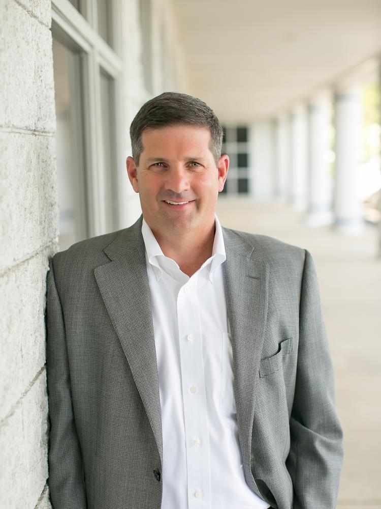 Photograph of Jimmy Hiller in a white shirt and gray blazer, leaning against a white brick wall with an outdoor background.
