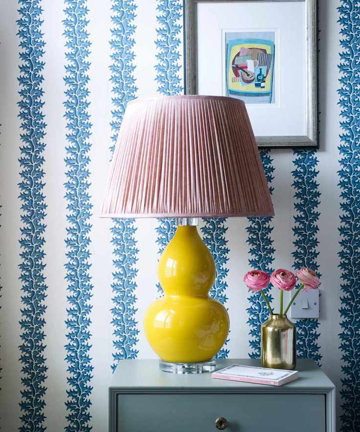 A room with blue and white statement wallpaper, a yellow ceramic lamp with pink lampshade, framed artwork in silver frame and blue bedside table with a gold vase filled with pink flowers and a book.