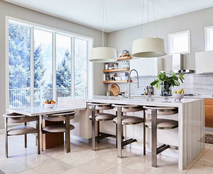 Kitchen with large, central island with different levels, area for bar seating and low area for dining seating, island crafted from natural stone, matching metal and dining chairs with upholstered seat and back cushions, two large pendant lights hanging over the island, white and wooden kitchen cabinets