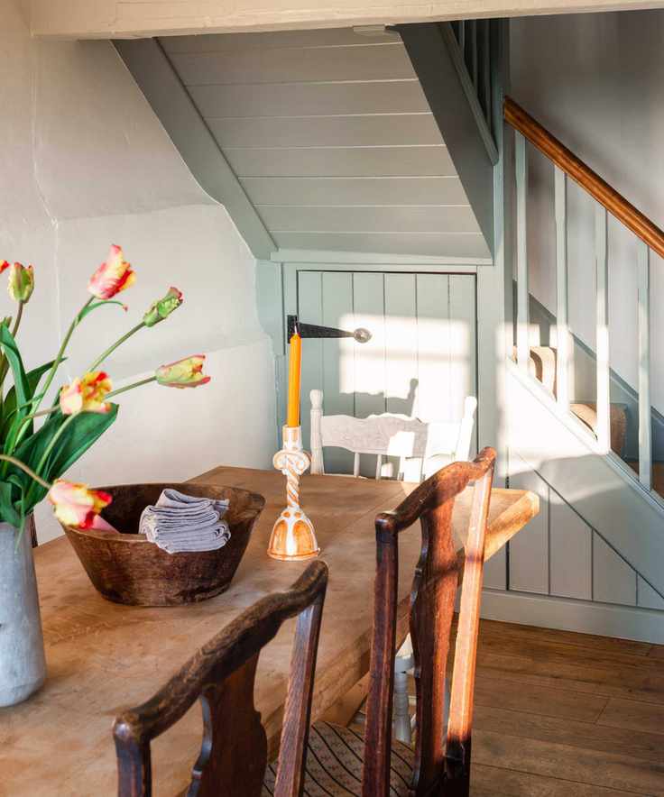 A cottage dining area with a wooden kitchen table, chairs and panelled staircase painted in a pale greenish gray paint. There is an orange candle, fruit bowl and vase of tulips on the table.