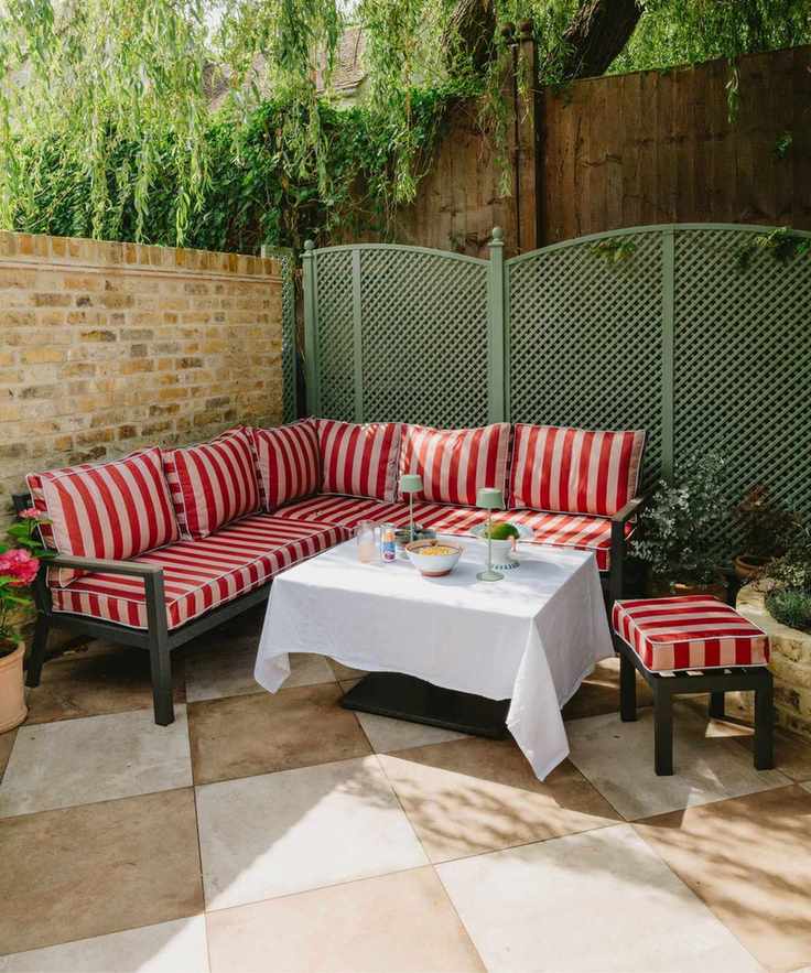 corner backyard patio with a brick wall, green painted trellis fence, beige and white checkerboard patio tiles, with a large corner sofa with pink striped cushions