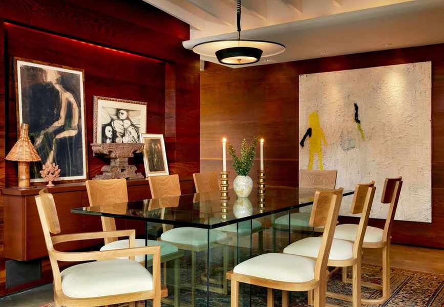 a dining room with a glass table, wooden dining chairs, wood paneled walls, and artwork hanging