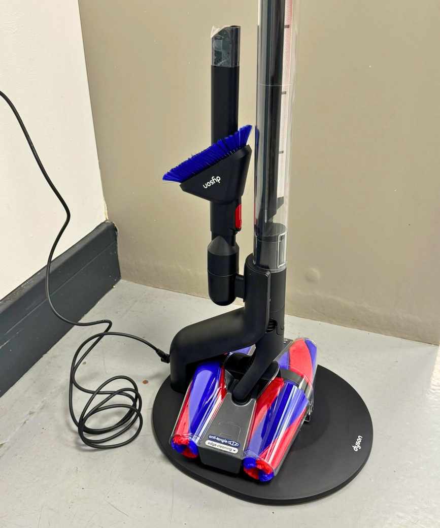 The Dyson PencilVac Fluffycones on its charging base, with attachments mounted, on a gray floor.