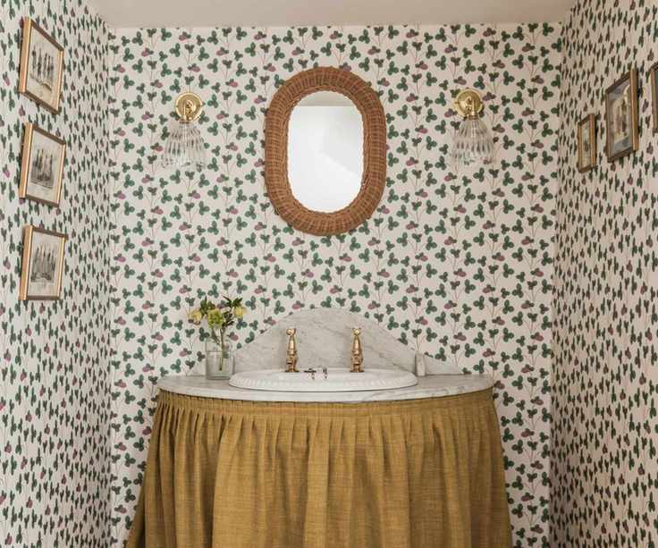 Vintage sink with upholstery curtain, marble basin and vintage wallpaper with rattan mirror, glass sconces and small framed pictures