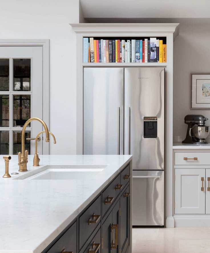 Close up of corner of kitchen with large double fridge in alcove, shelving and cabinetry surrounding, kitchen island with sink, brass tap