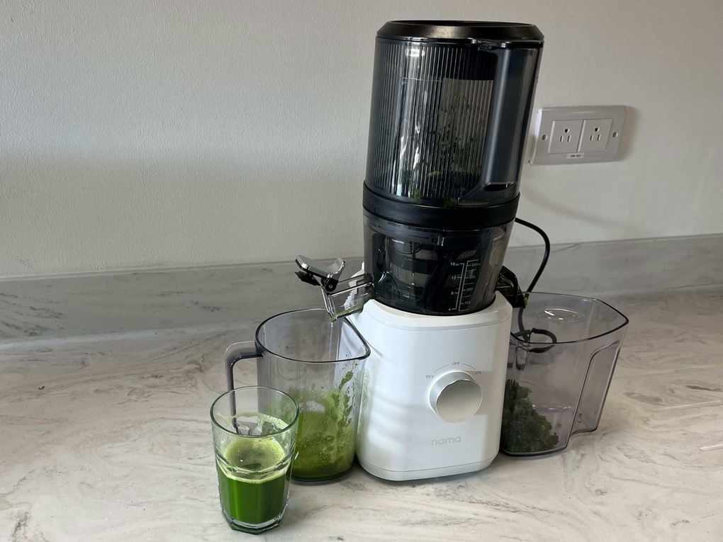 A cold press juicer with a white plastic base and a black plastic top, dispensing green juice into a beaker on a marble kitchen countertop.
