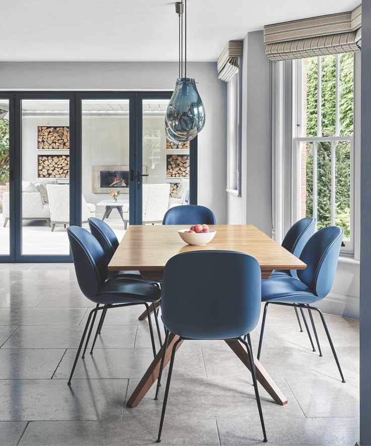 A bright dining room with a wooden table and blue chairs, with a fruit bowl on top, tiled gray flooring, and a white fruit bowl on top of the table. Above the table are blue glass pendant lights, and to the right are bay windows with striped blinds. In the background are glass doors out onto a patio with a white wall, lit wood burner, log store, and white chairs.
