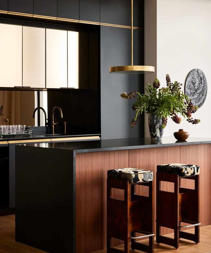 Black and dark wood kitchen with gold accents. A golden light hangs above a kitchen island next to two stools, with a gold faucet and black and reflective cabinets pictured in background.
