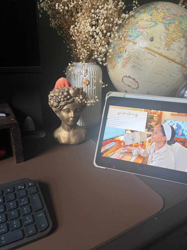 An ipad with a youtube video on, on a brown office desk, in front of an old globe, dried stems, and a gold bust with a cleaning cloth inside