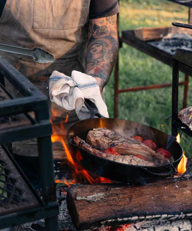 A vintage-style cast iron frying pan being used over lit logs to cook steaks.
