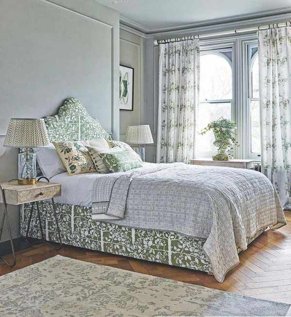 A pale green bedroom space with sage panelled walls and a double bed with a green pattern, surrounded by patterned bedside tables and lamps. The floor is light wood, featuring a large patterned rug. In the background, two arched windows with white and green floral-patterned curtains allow natural light to flood the room.
