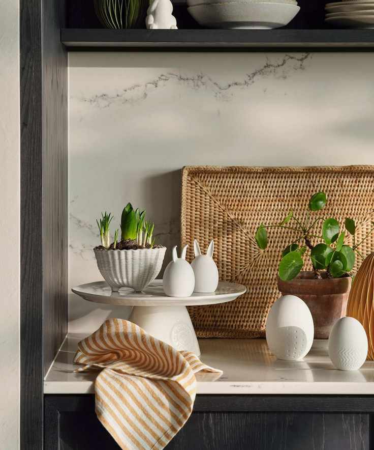 A white kitchen counter with Easter decorations on a cake tray, beside some freshly sprouting plants and flowers.