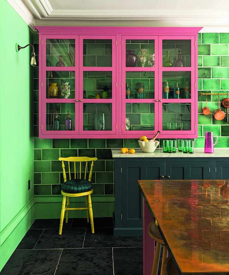 An example of small kitchen storage ideas showing pink, glass-fronted cabinets in front of a green-tiled wall and above a yellow chair