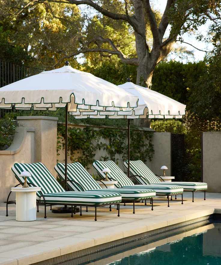 a poolside set up with four lounge chairs with striped cushions, two umbrellas with green trim, and side tables with lamps on each one