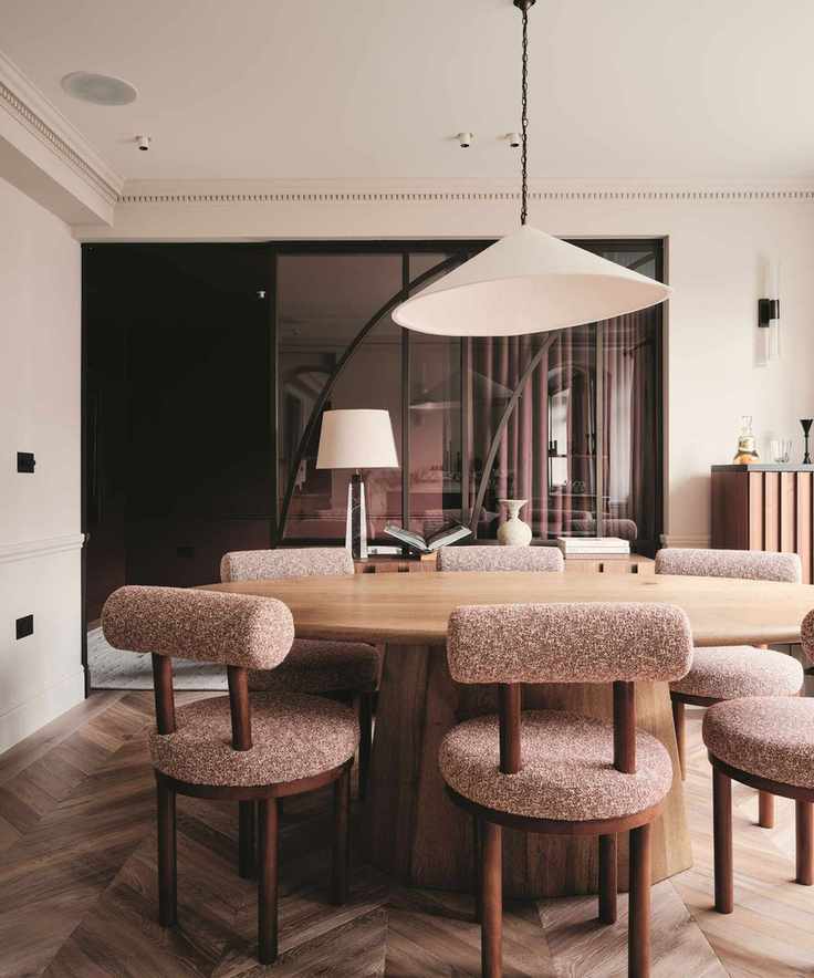 Oval oak dining table with mauve bouclé chairs under a large conical pendant. A curved glass partition and chevron wood floors complete the modern, warm interior.