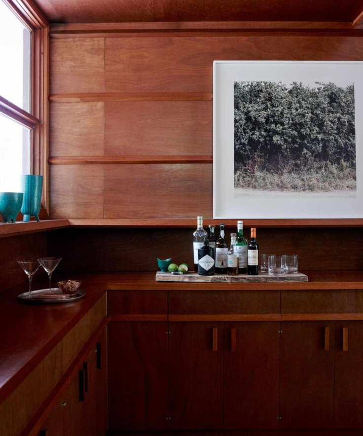 Midcentury bar with dark wood panelling, blue glasses on window sill and framed photograph, below are alcohol bottles and glasses