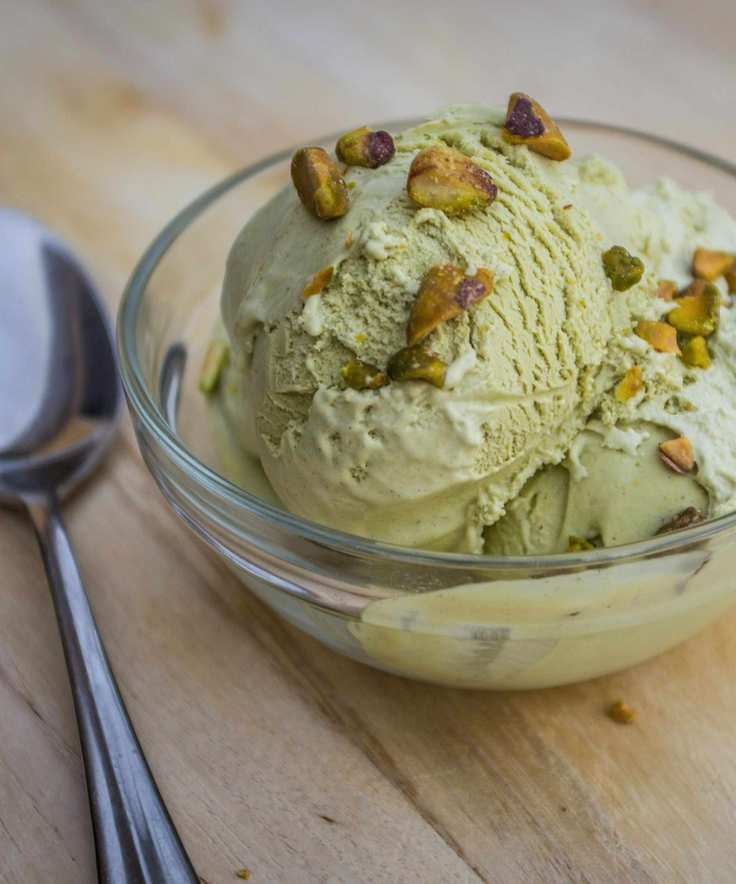 Glass bowl of pistachio ice cream with crushed pistachios on top, placed on a wooden table with a silver spoon beside it