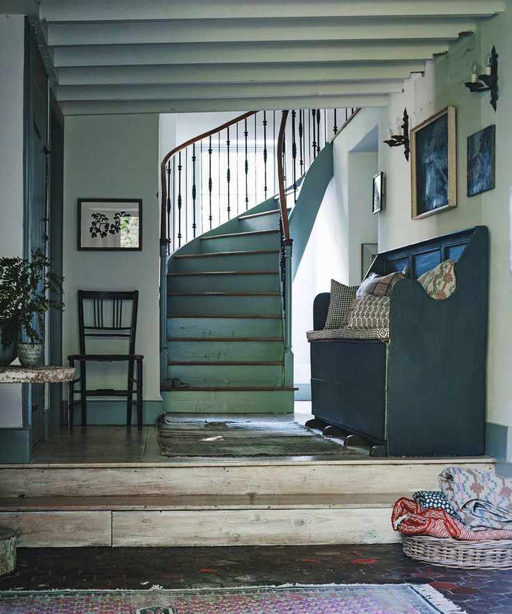 A blue, farmhouse style entryway with light worn wooden steps and panelled floor up to a curved blue staircase with a wooden banister. To the right is a wooden bench seat with patterned cushions below framed artwork, and to the left is a wooden chair.
