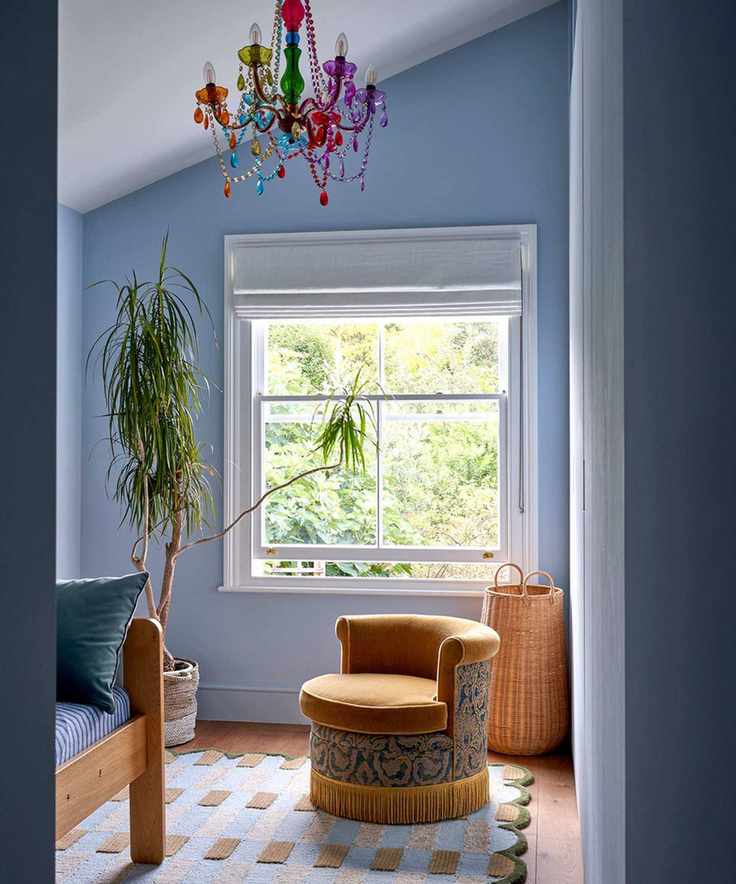A bedroom with powder blue walls, white ceiling and window trim, a blue patterned rug, a wooden bed, and a small ochre chair.