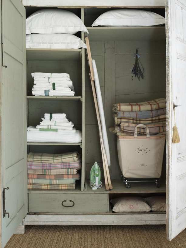A green painted linen closet with rustic bed linens folded on shelves beside a leaning ironing board.