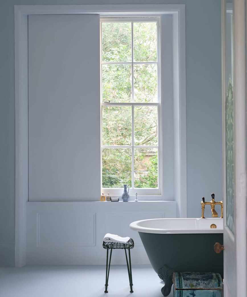 A light and airy bathroom with pale blue walls and a large window, and a dark blue roll-top bath.
