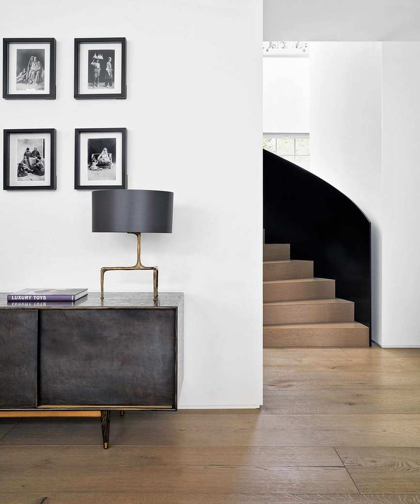 Curved wooden staircase into room with wooden floor and dark wood floor. White painted walls, dark gray, metallic sideboard, table lamp with dark gray shade, four framed images in black and white on wall above sideboard