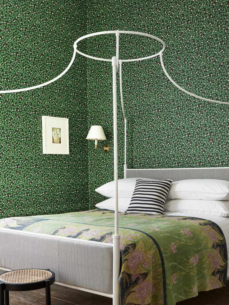 green bedroom with ditsty green floral wallpaper, white iron bed, white artwork, white wall light, stool, black and white stripe cushion, green, pink and yellow floral blanket