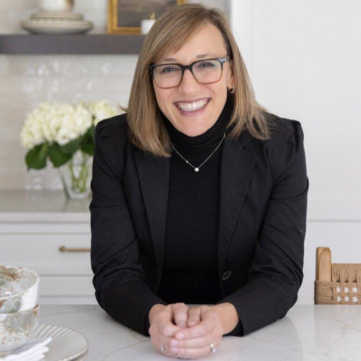a photo of interior designer Elana Mendelson leanin on a kitchen countertop: a lady in all black with black glasses and light brown bob hair