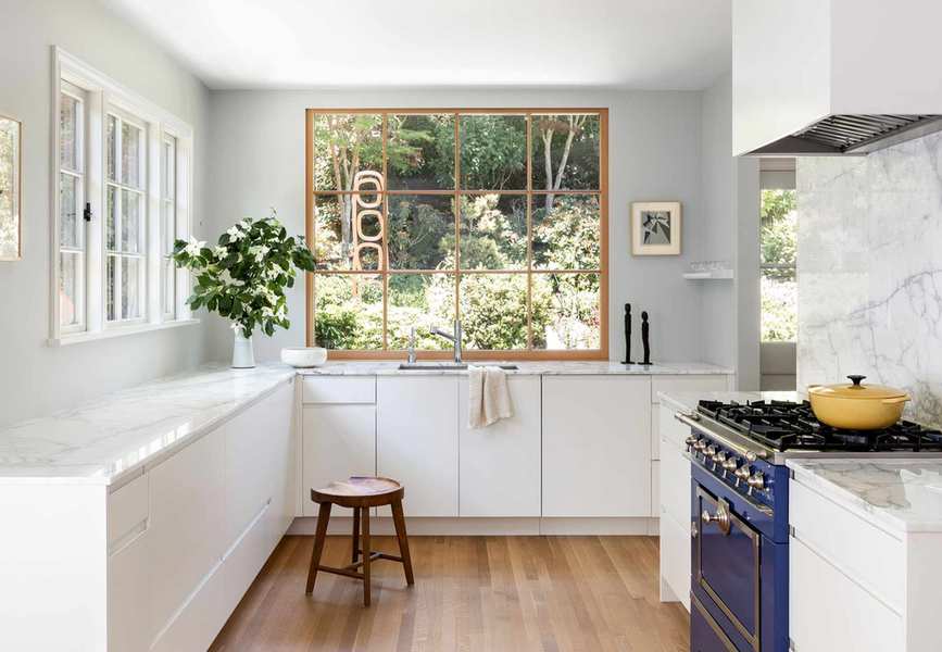 A kitchen showcasing large windows, white lower cabinets, and a blue La Canche stove, illustrating white kitchen backsplash inspirations.