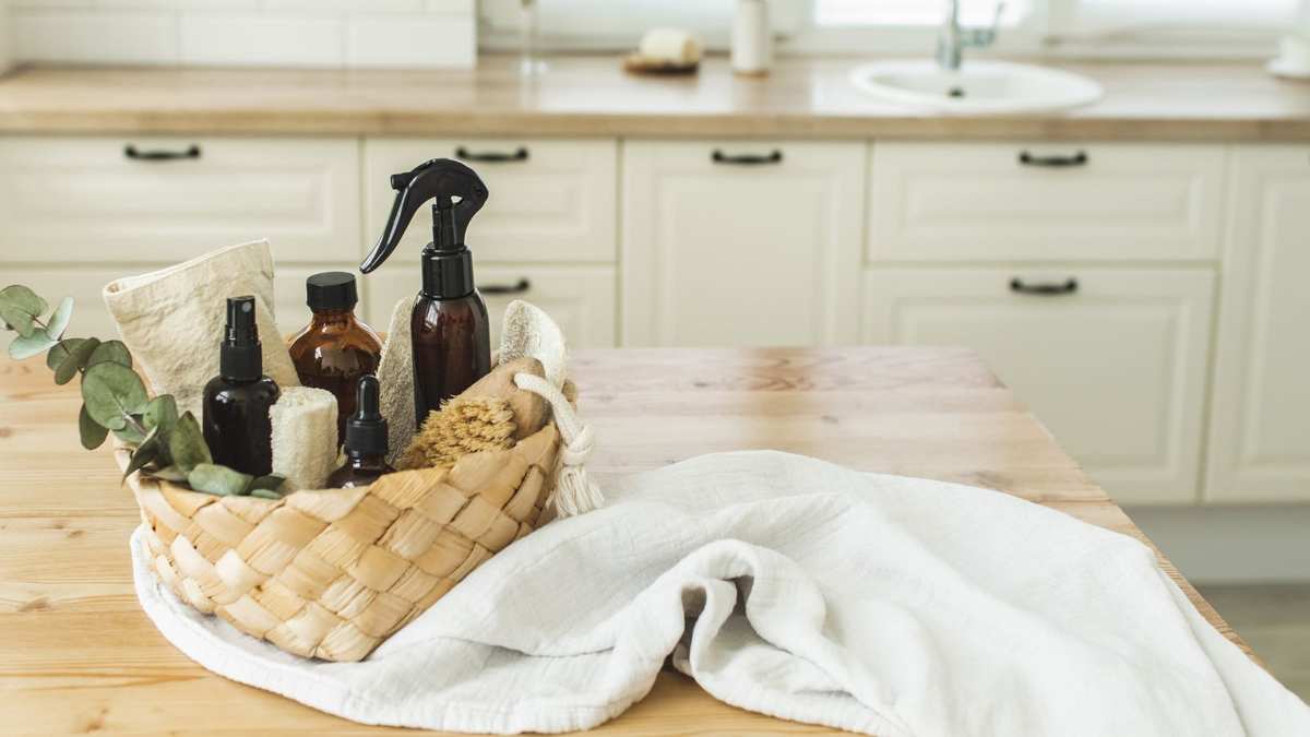A woven basket of amber glass cleaning bottles and a white rag on a wooden kitchen counter top, blurry white cabinets and a kitchen sink in the background