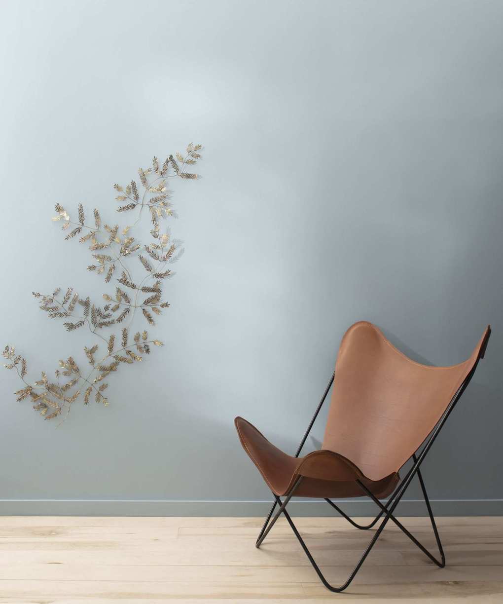 A wall painted in a cool grey paint, with a decorative wall hanging reminiscent of a branch with leaves. A leather chair is in front of the wall.