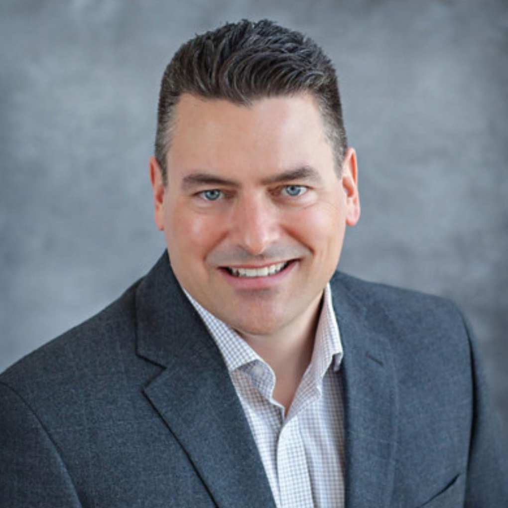 A picture of Ron Shimek - a man wearing a gray blazer and checked shirt, with a gray background behind him