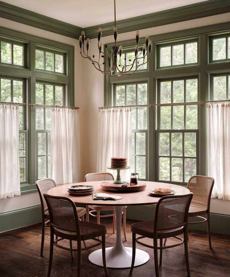 A breakfast nook in a kitchen with large windows with green woodwork and cafe curtains, wood floors, a round dining table, and wooden chairs.